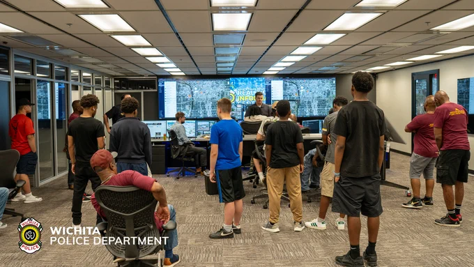 A group of people stands inside a modern control room with multiple screens displaying maps and data. The room is part of the Wichita Police Department. Individuals are engaged in observing and discussing the information displayed.
