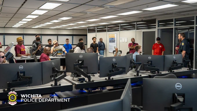 A group of people are gathered in a room with multiple computer stations and monitors, appearing to participate in a presentation or discussion. Wichita Police Department branding is visible.