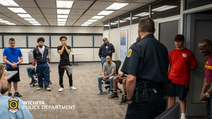 A group of people is gathered in a room with office-style lighting. They are standing or sitting, listening to a person in a police uniform speaking. The Wichita Police Department logo is visible on the image.