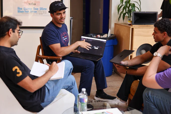 A group of people in a casual meeting setting. One person is holding a laptop, which displays a website, while speaking to others. They are seated in a room with a framed document, a recycling bin, and a speaker. Some participants are taking notes or holding a device.