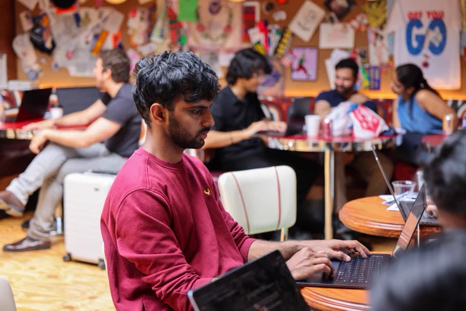 A person intently works on a laptop in a lively cafe-like setting, surrounded by others engaged in conversation and using devices. The walls are decorated with various colorful posters and artwork.