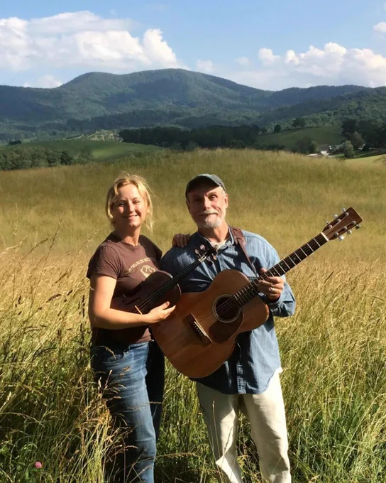 Two people standing in a sunlit grassy field holding acoustic guitars with rolling green hills and a blue sky in the background.
