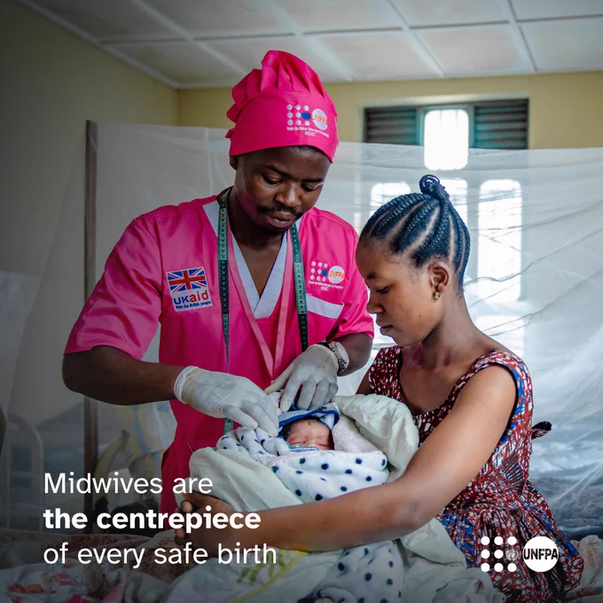 A photo of a midwife in pink medical scrubs with the UNFPA logo on them standing next to a seated woman who holds a baby in her arms, swaddled in a white blanket. White text overlaid on the photo reads “Midwives are the centrepiece of every safe birth.” The UNFPA logo is in the bottom right corner.