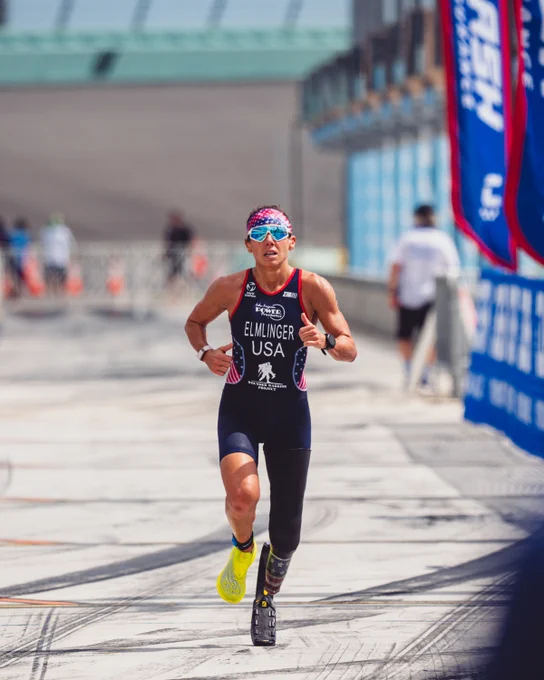 Athlete with a prosthetic leg running in a race, wearing a USA uniform and sunglasses, with banners and spectators in the background.