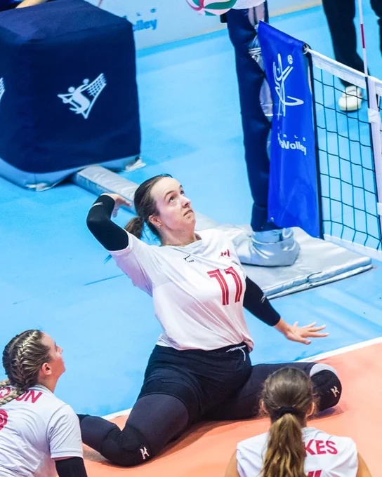 A seated volleyball player in a white jersey reaches for the ball near the net, surrounded by teammates on a blue court.