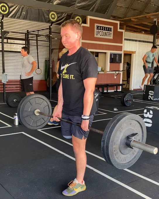 Person lifting a barbell in a gym, wearing a black shirt and shorts. Others exercise in the background.