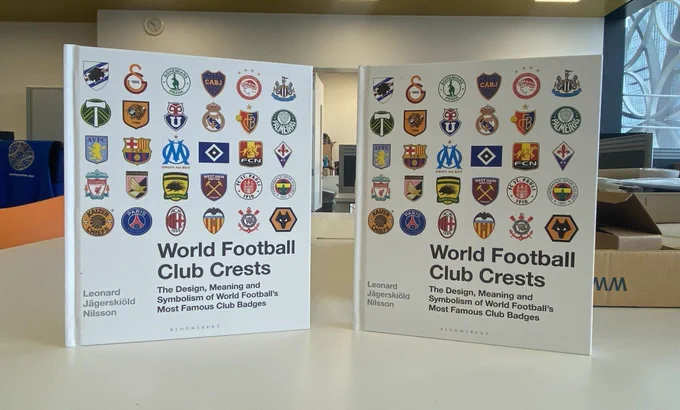 Two copies of a book on a table -  covers facing toward the camera. Title of the book on the bottom right of the front cover is World Football Club Crests. The author’s name on the bottom left of the cover. Above this are rows of football club badges. An office scene behind the table on which the books are standing. A desk on the left with a waistcoat on the back of a chair. An open doorway in the background between the two books. A window on the right of the room.