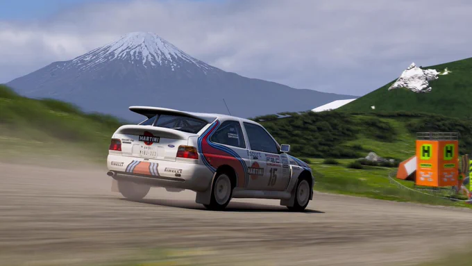 A rally car, adorned with Martini Racing livery, speeds along a dirt road with a snow-capped mountain in the background. Green hills and distant structures with bright orange-and-green signs are visible on the right.