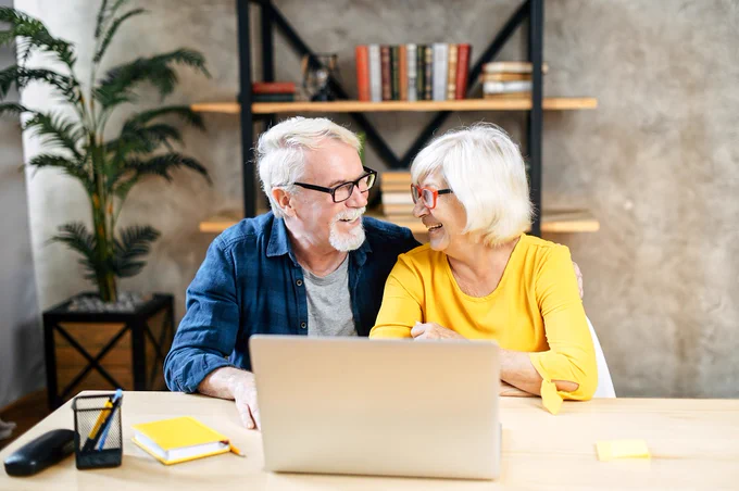 Senior couple sitting at a wooden table using a laptop together in a cozy, modern living room with bookshelves and plants.