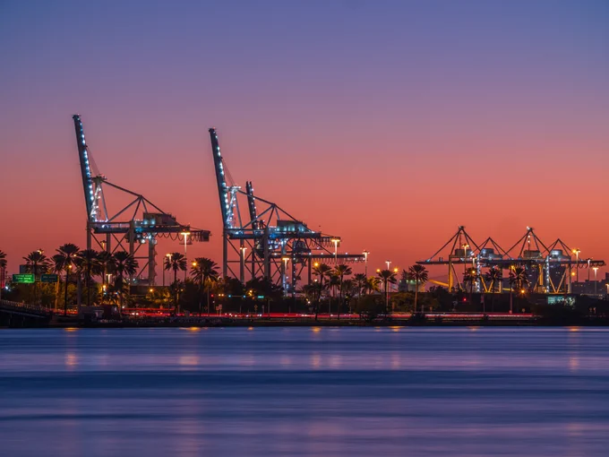 Industrial port at dusk with a gradient sky from purple to orange. Several large, illuminated cranes stand tall behind palm trees and a highway with streaking car lights. Water in the foreground reflects the port's lights. Signs read: 'Terminal Island Fisher Island NEXT LEFT', 'Star Island NEXT RIGHT'.