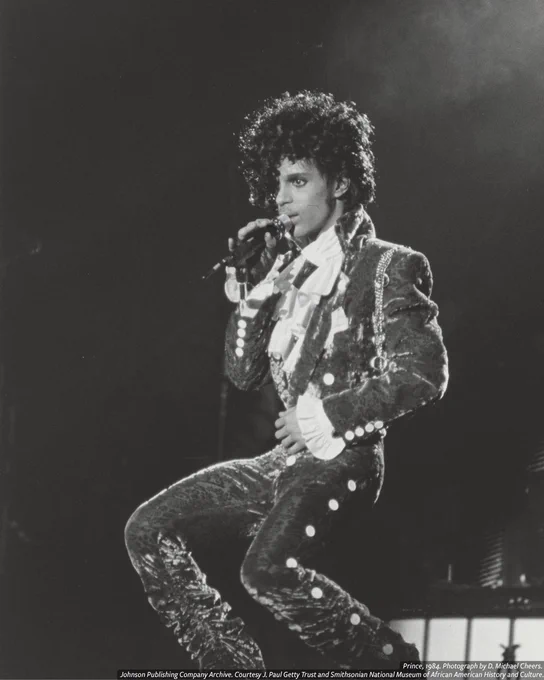 Black-and-white photograph of Prince performing, singing into a microphone while wearing a stylish outfit featuring lush fabric, ruffles, and custom accessories. At the bottom is the credit line, ‘Prince, 1984. Photograph by D. Michael Cheers. Johnson Publishing Company Archive. Courtesy J. Paul Getty Trust and Smithsonian National Museum of African American History and Culture.’ 