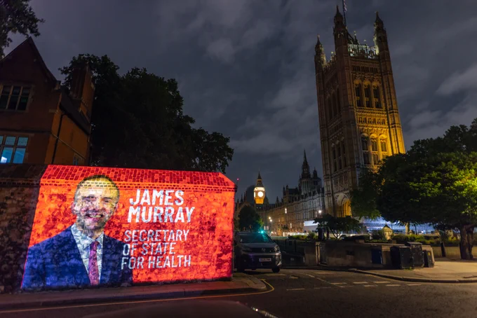 A photograph of James Murray Secretary of State for Health (white text on red background) is projected onto a wall wit the Houses of Parliament in the background at night.