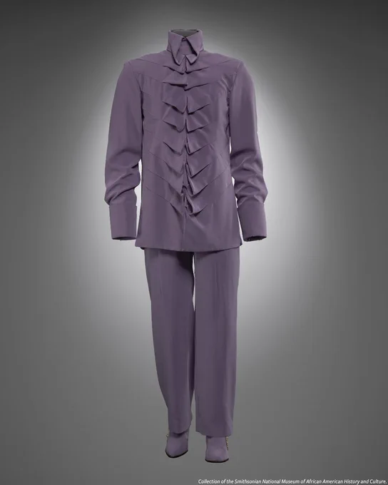 Prince's lavender satin outfit, featuring a flowy long-sleeved shirt, high-waisted pants, and matching boots with custom zippers. At the bottom is the credit line, ‘Collection of the Smithsonian National Museum of African American History and Culture.’ 