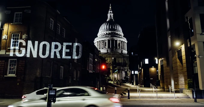 The word Ignored is projected onto a building in London with St Paul's Catherdral in the background lit up at night.