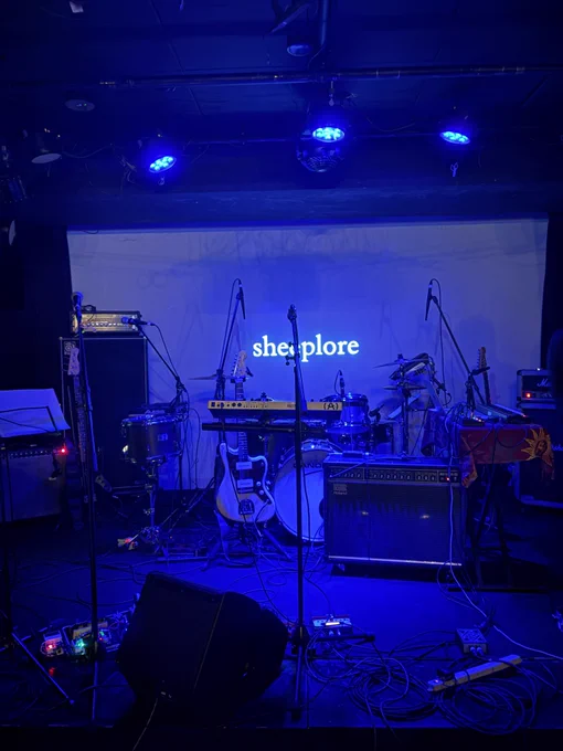 青い照明に包まれた神楽音のステージ。中央にはドラムセットが置かれ、ギターやキーボード、複数のマイクスタンドやアンプ、エフェクターボード、ケーブル等がステージ上に並んでいる。背面のスクリーンには「sheeplore」の文字が白く投影されている。