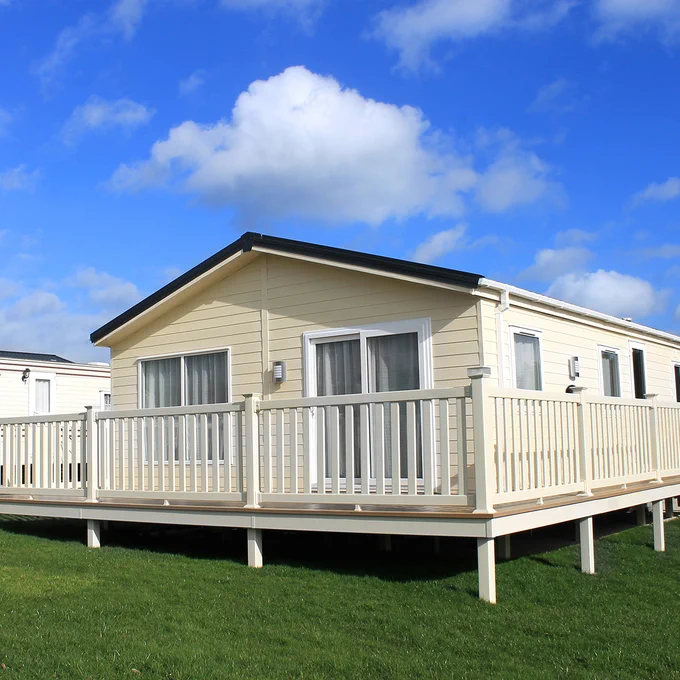 A beige mobile home with white trim and a wooden deck is set on a grassy area under a partly cloudy sky.
