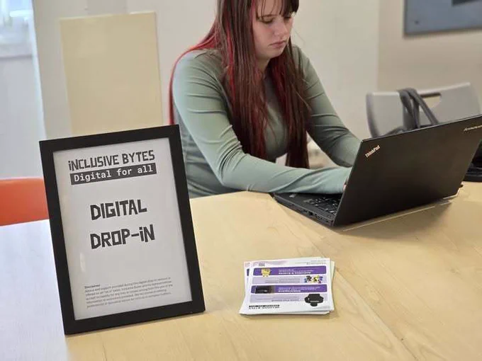 A young person sat behind a sign and a laptop, the sign reads "Inclusive Bytes Digital For All - Digital Drop-In"