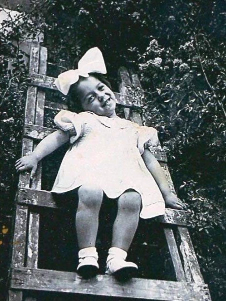 A young girl with a large bow in their hair sitting joyfully on a wooden ladder, in a vintage black-and-white photograph.
