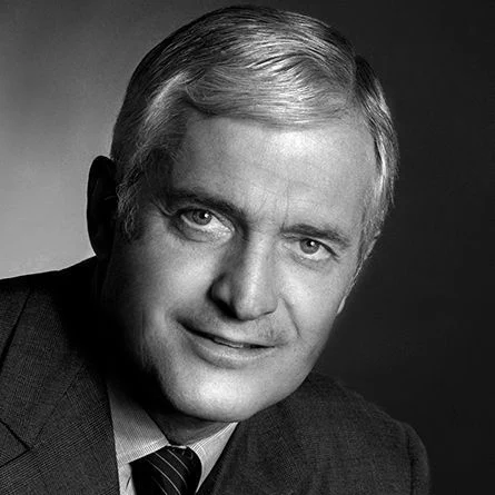 John Tuner in a black-and-white studio portrait, wearing a suit jacket, dress shirt and striped tie. He has light-coloured hair combed to the side and faces the camera with a slight smile. The background fades from light gray on the left to dark gray on the right.
