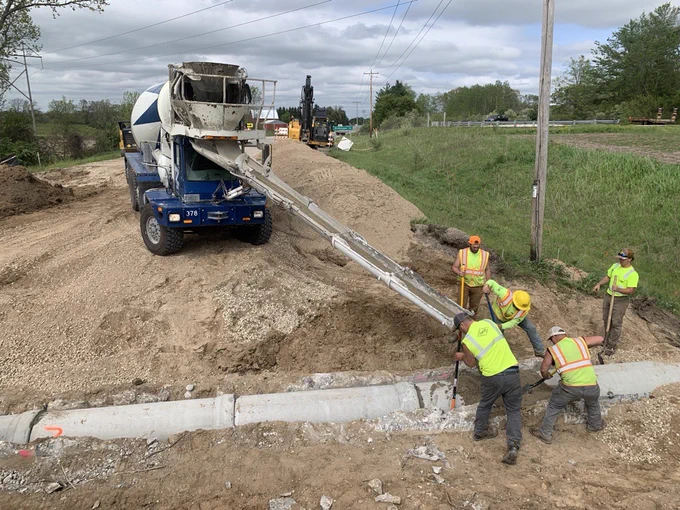 Construction workers in safety vests and helmets installing concrete pipes with a cement mixer truck pouring concrete on a rural roadside.