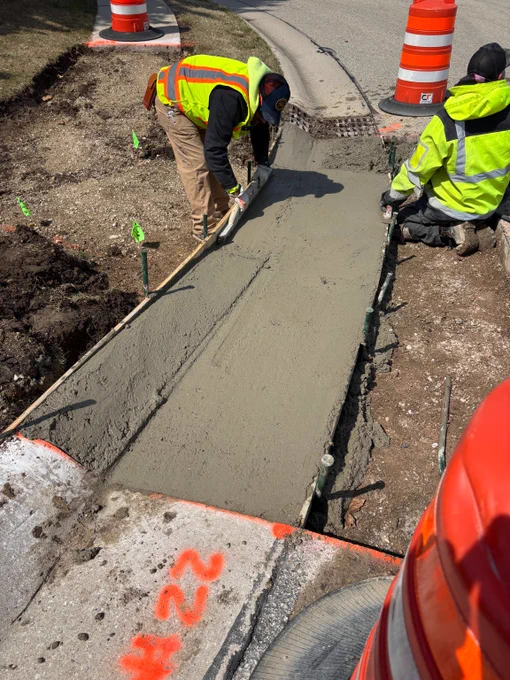 Two construction workers smoothing freshly poured concrete on a sidewalk section beside traffic barrels.