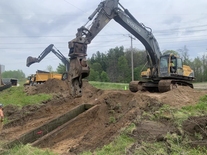 Excavator with hydraulic hammer attachment breaking ground at a construction site with dirt piles and trees in the background.