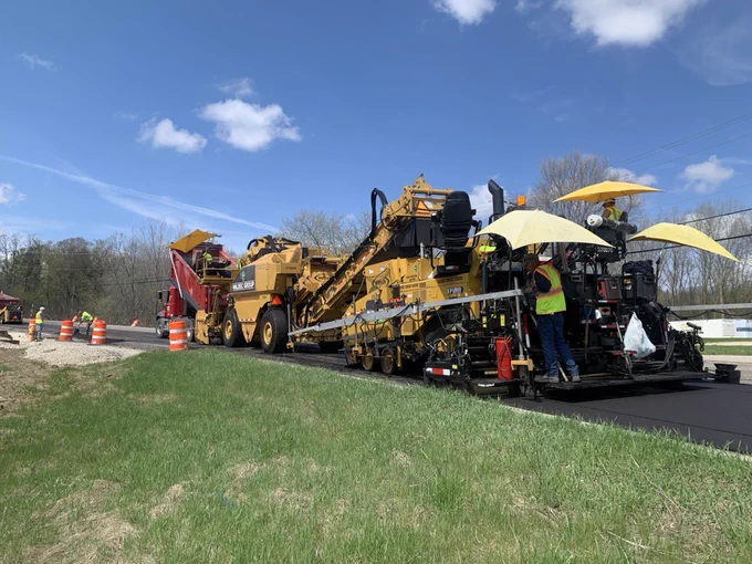 Construction workers operating heavy machinery to lay fresh asphalt on a rural road under a clear sky.