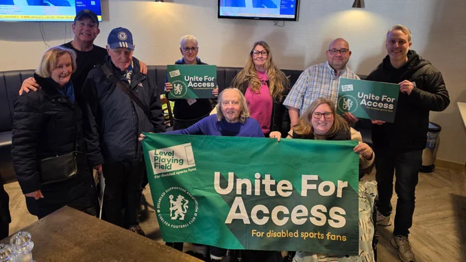 A group of people, smiling towards camera and holding up green 'Unite For Access' boards and a flag, which all feature the Chelsea Disabled Supporters' Association logo.
