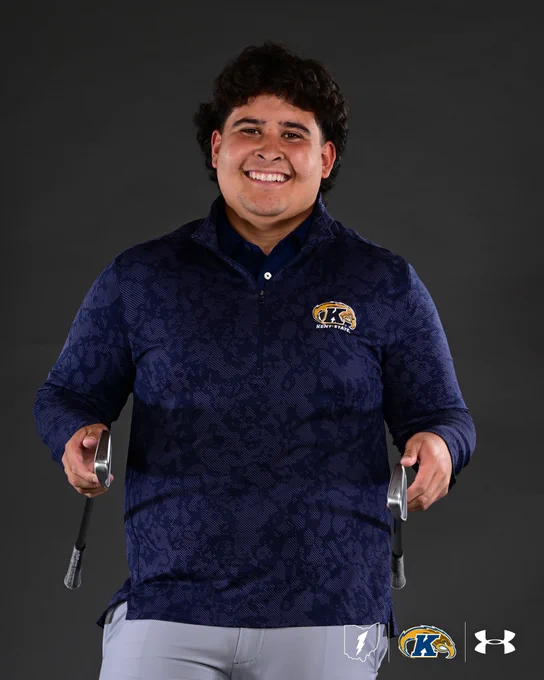 An official portrait photo of Kent State men's golf player Isaiah Ibit smiling broadly against a dark gray studio background. He is wearing a navy blue Kent State quarter-zip pullover with a subtle textured pattern throughout, featuring the Kent State Golden Flashes logo embroidered on the left chest, layered over a navy collared polo shirt. He is paired with light gray golf pants. Isaiah is holding a 6 iron in his right hand and a 7 iron in his left hand, both at his sides, in celebration of 67 Day. He has short dark curly hair and is looking upward with a wide smile. The Ohio state outline with lightning bolt, Kent State Golden Flashes logo, and Under Armour logo are displayed in the bottom right corner of the image