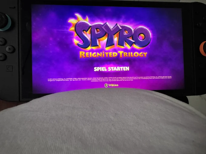 Eine tragbare Spielekonsole zeigt den Startbildschirm von Spyro Reignited Trilogy. Der Bildschirm leuchtet in kräftigen Violett- und Pinktönen, während die Konsole auf dem Schoß oder Bauch einer liegenden Person ruht. Die Szene vermittelt einen entspannten Gaming-Moment vor dem Start eines Abenteuers.