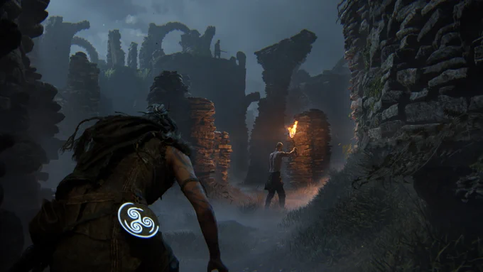 Senua, a Celtic warrior, sneaks between stone ruins. Two humanoid creatures prowl in the shadows. One wields a flaming torch.