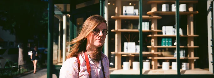 mb, a white blonde trans girl, stands in front of a shop window, hair blowing in the breeze, wearing round glasses and a pink Paper Plane patterned shirt and red camera strap.