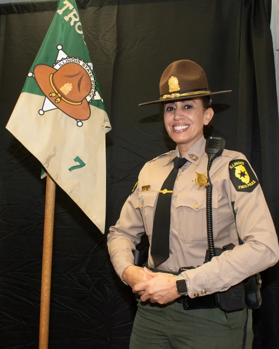 A person in a law enforcement uniform stands smiling in front of a flag displaying the Illinois State Police logo. The uniform includes a brown hat and various insignia, with a visible badge and Illinois State Police shoulder patches. The background features a dark curtain.