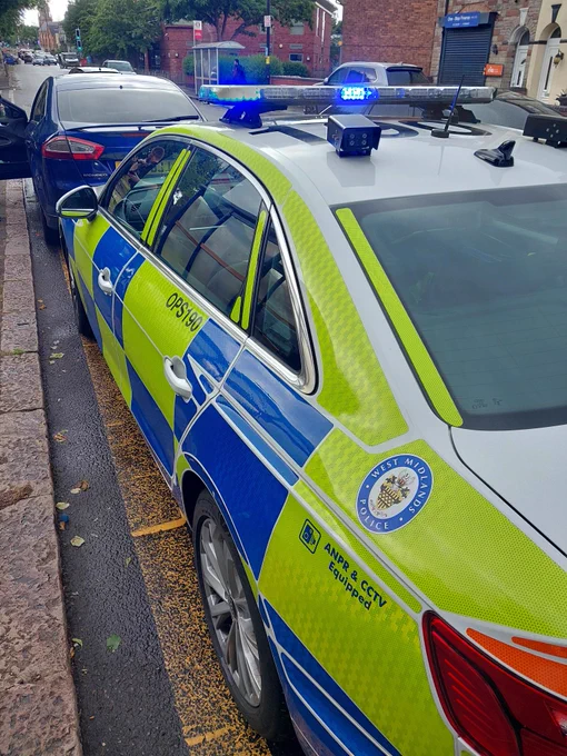 West Midlands Police car with blue lights flashing, parked on a street next to another car.