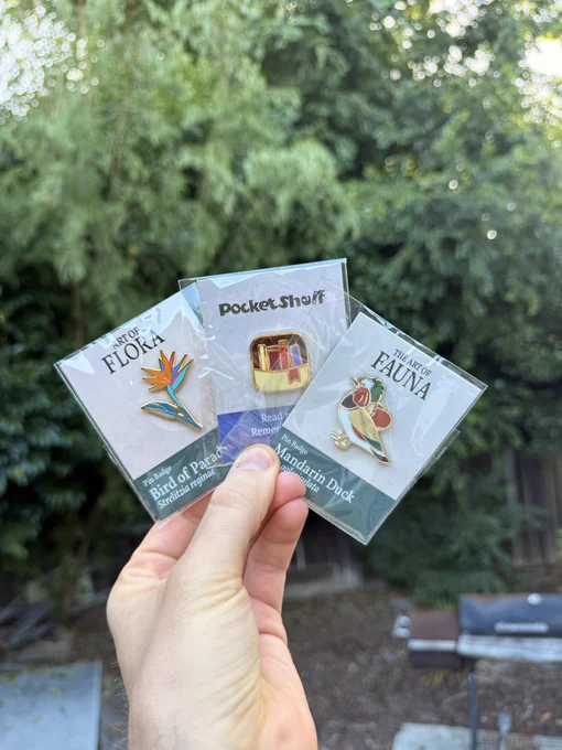 Me holding three pins in plastic package. It’s Art of Flora, PocketShelf and Art of Fauna.