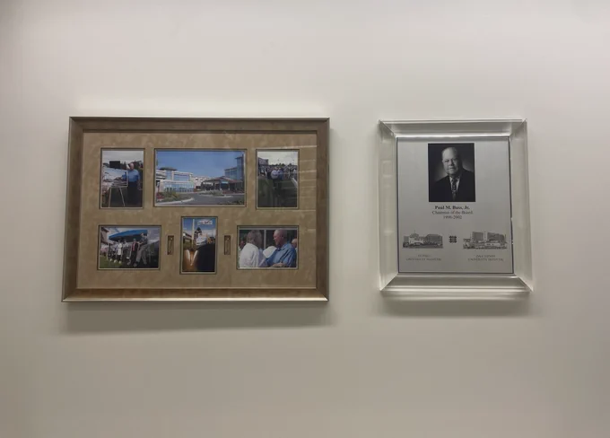 Two items hanging on a white wall. The one on the left is a framed collage of six photos and two golden shovels, taken at the ground breaking ceremony for the Bass Center. On the right is a glass mounted plaque that reads "Paul M. Bass, Jr. Chairman of the Board 1998 - 2002 St. Paul University Hospital / Zale Lipshy University Hospital".