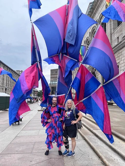 A medium shot of Robyn Ochs standing outside on a plaza next to a person wearing a massive, elaborate flag costume. Robyn is on the right, smiling with her hand on her hip, wearing a dark blue t-shirt, black shorts, and blue sneakers. The person on the left is wearing a ruffled outfit in pink, purple, and blue, standing in front of a giant, towering structure of multiple bisexual pride flags fanning out around them like a cape.