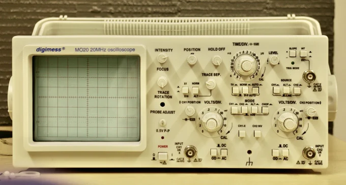 Photo of a bench oscilloscope angled from the front-left, with a green waveform trace visible on the screen and control knobs across the front panel.