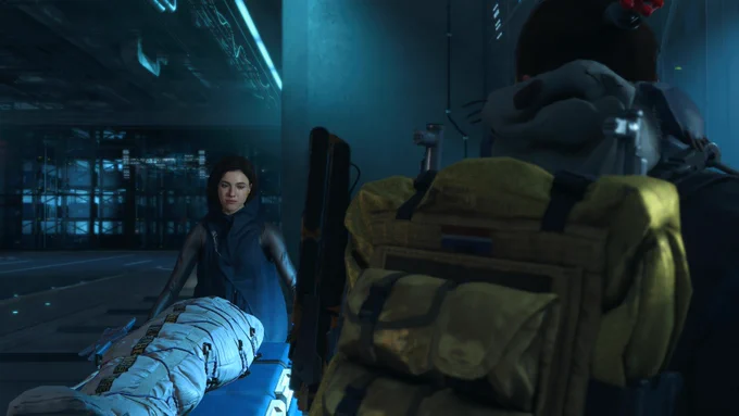 Sam liefert in Death Stranding Directors Cut eine verpackte Person auf einer blauen Liege in einer Station ab. Eine Frau in dunkler Kleidung nimmt die Lieferung im kühlen Licht entgegen.