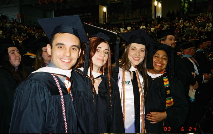 A group of graduates in caps and gowns smile at the camera at a graduation ceremony. In the background, a large crowd of people is seated in an indoor arena, attending the event.