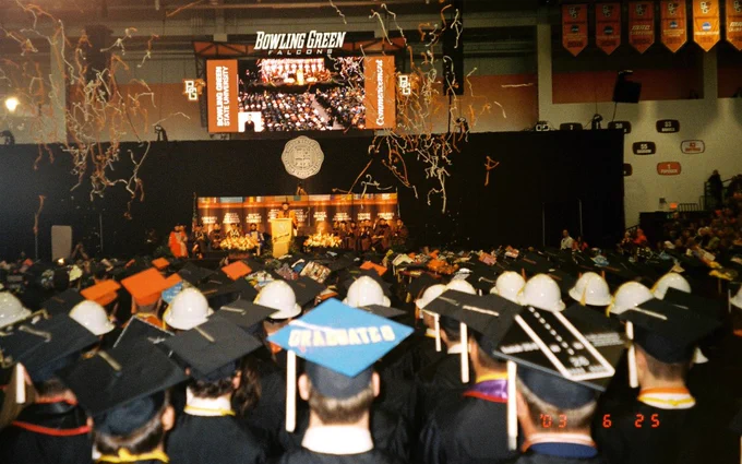 A graduation ceremony at Bowling Green State University. Graduates in caps and gowns face a stage with a large emblem and banners. Streamers fall from above, and a large screen displays the event.