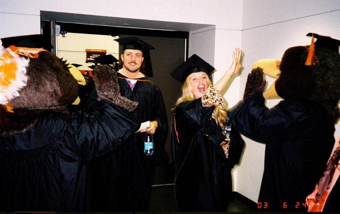 Graduates in caps and gowns gather in a hallway, some interacting with costumed mascots of Bowling Green State University. A large screen in a gymnasium displays "Bowling Green" as the graduates embrace in a circle.
