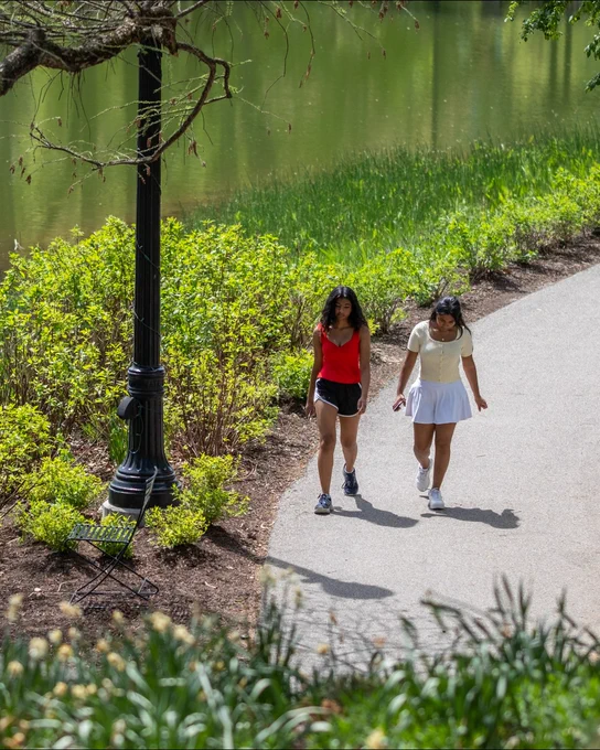 Two people are walking along a paved path beside Mirror Lake. They are dressed in casual athletic wear and are walking near green foliage and a lamppost with an empty chair beside it.