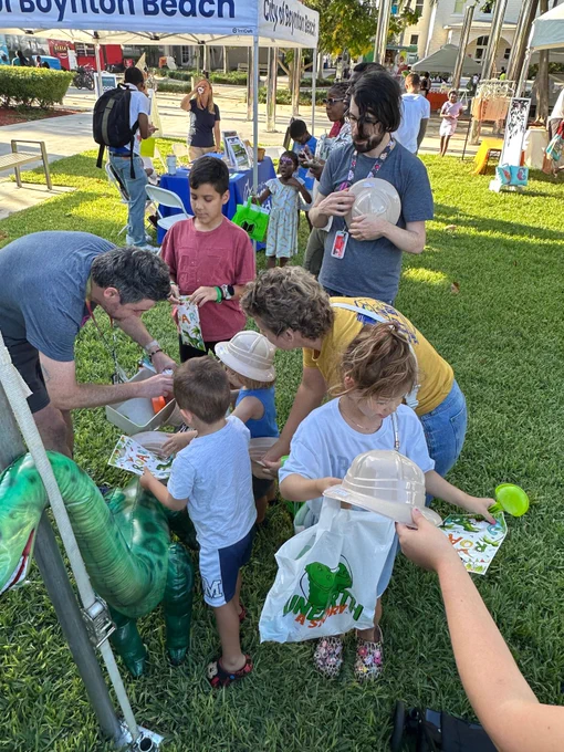 Children and adults gathered outdoors on a sunny day at a City of Boynton Beach event, engaging with educational materials and dinosaur-themed activities on a grassy area with tents and displays in the background.