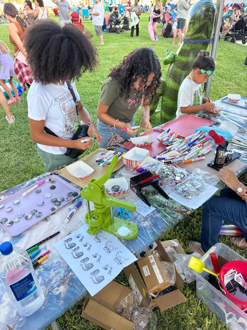 Children and a young adult engaged in a button-making craft activity at an outdoor event, surrounded by colorful markers, button parts, and a green button press machine on a table covered with supplies and instructions, with other attendees visible in the background on a grassy area.
