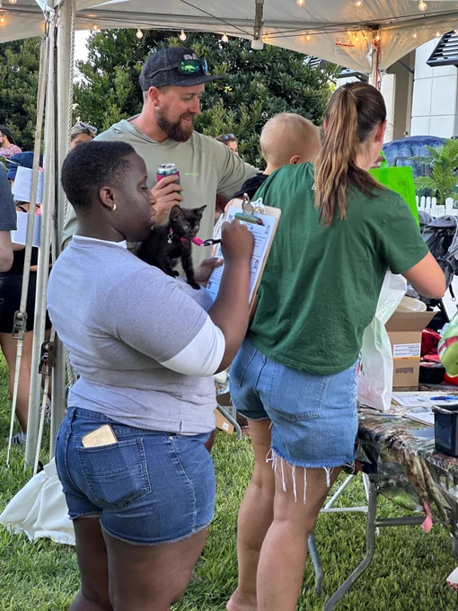 A group of people gathered under a tent at an outdoor event, with a young person holding a clipboard and a small black cat, a man holding a baby, and a woman organizing items on a table covered with various supplies.