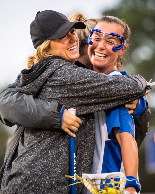 A lacrosse player in a blue and white uniform is being warmly hugged by a person in a gray jacket and black cap, both smiling and expressing joy.