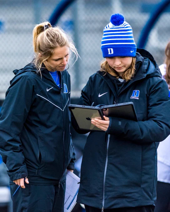 Two women in sports jackets are reviewing a digital tablet on a sports field, possibly discussing a game plan.