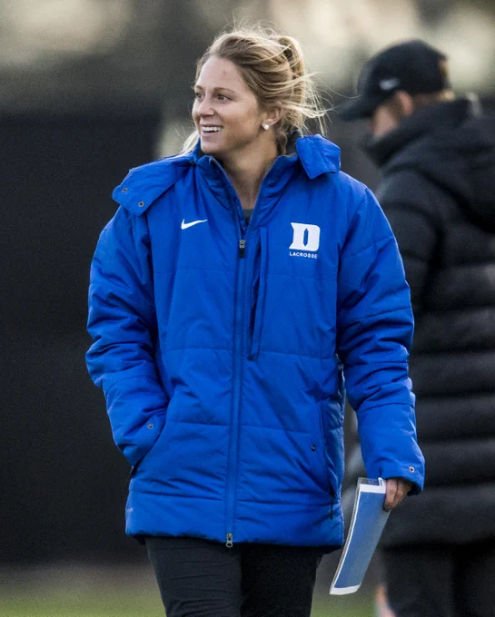A woman in a blue jacket with a 'D' logo is smiling, holding a clipboard. She stands outdoors on a field, with another person in a dark jacket slightly blurred in the background.