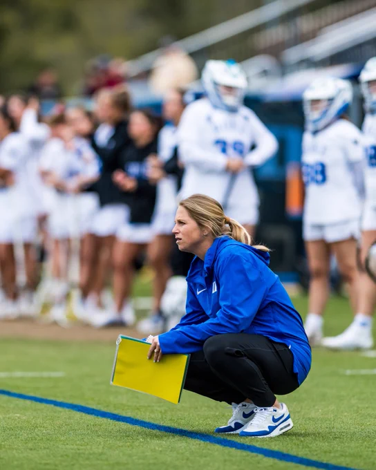 A coach in a blue jacket squats at the edge of a sports field, holding a yellow clipboard, with a group of players in white uniforms standing blurred in the background.